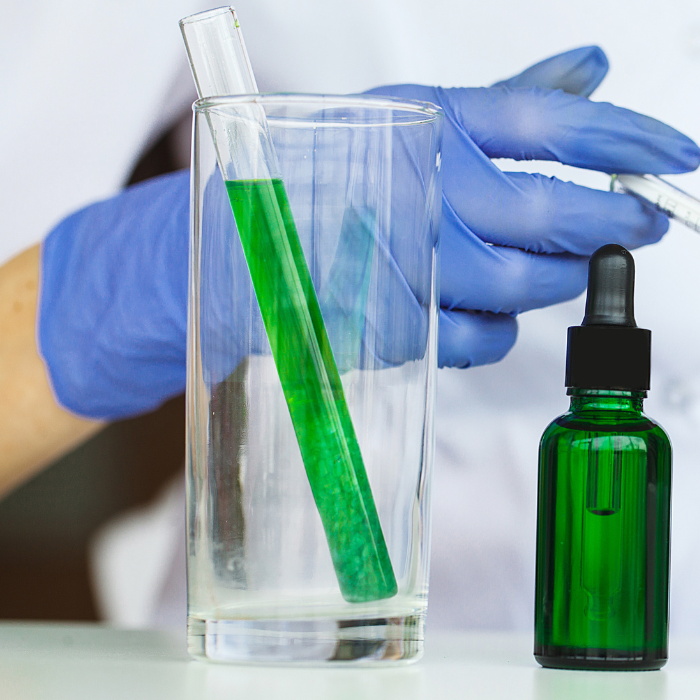green testtube in a glass with a green serum jar next to it. A gloved hand in the background holds a pipette